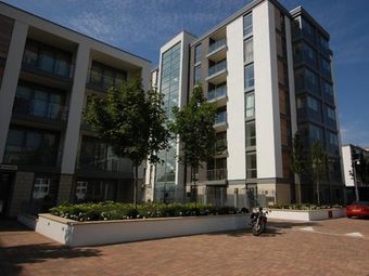 Modern multi-story apartment buildings with balconies, surrounded by trees and landscaped planters. A motorcycle and car are parked on the paved area.