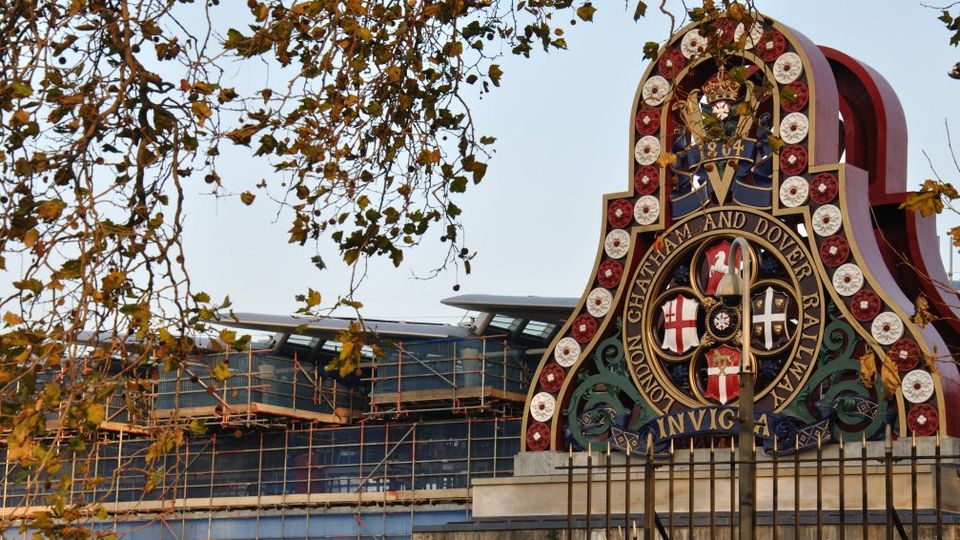 Ornate railway emblem with historical symbols and "London Chatham and Dover Railway" text, framed by autumn branches against a building backdrop.