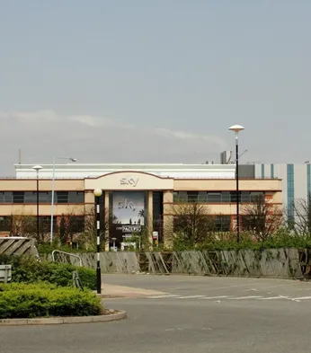 A large office building with "Sky" signage, surrounded by trees and a parking area, set against a clear sky.