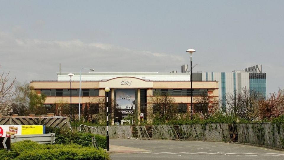 A large office building with "Sky" signage, surrounded by trees and a parking area, set against a clear sky.