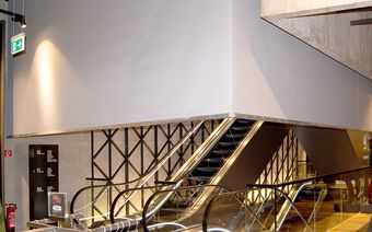 Modern indoor escalator with metal railings and wall lighting in a spacious building. Exit sign and directory visible on the left.