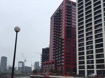 Tall red and white residential buildings in London, situated near a river. Cranes and other buildings visible in the background. The sky is overcast.