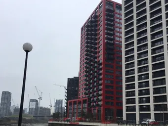 Tall red and white residential buildings in London, situated near a river. Cranes and other buildings visible in the background. The sky is overcast.