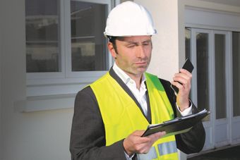 A man in a hard hat and yellow vest holding a phone and clipboard stands outside a building with a thoughtful expression.