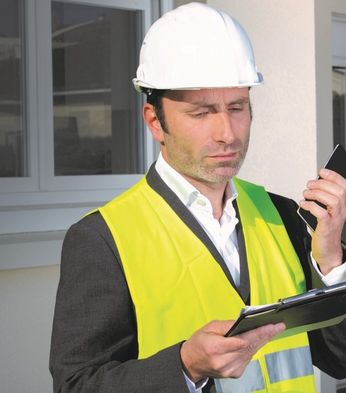 A man in a hard hat and yellow vest holding a phone and clipboard stands outside a building with a thoughtful expression.