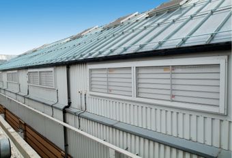 A corrugated metal building with a glass roof, featuring natural smoke shafts and window actuators, and a clear blue sky above.