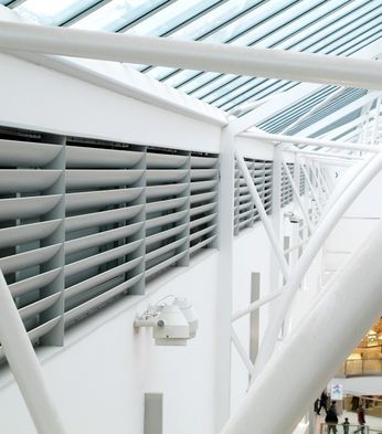 Interior view of a modern shopping centre, showcasing structural beams and louvered panels, with a glimpse of the shops below.