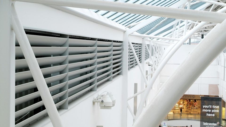 Interior view of a modern shopping centre, showcasing structural beams and louvered panels, with a glimpse of the shops below.