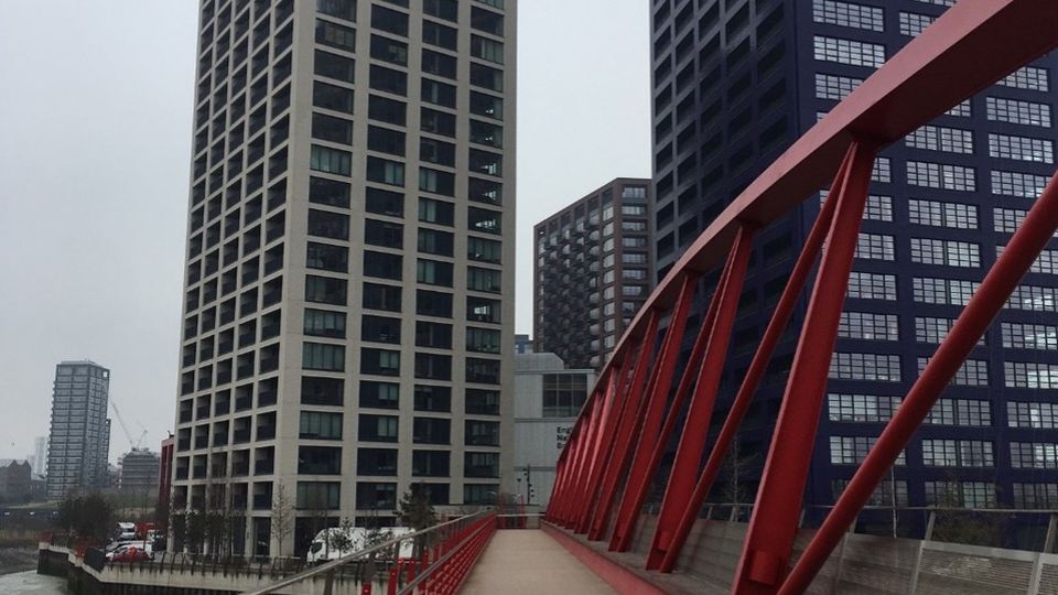 Red pedestrian bridge leading to tall modern residential buildings in London, under an overcast sky.