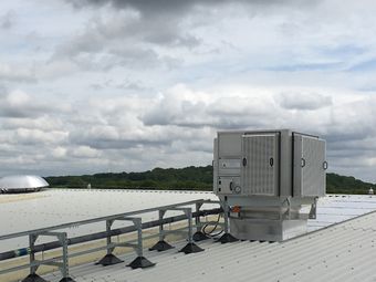 A rooftop air conditioning unit on a corrugated metal roof under a cloudy sky, with trees visible in the background.