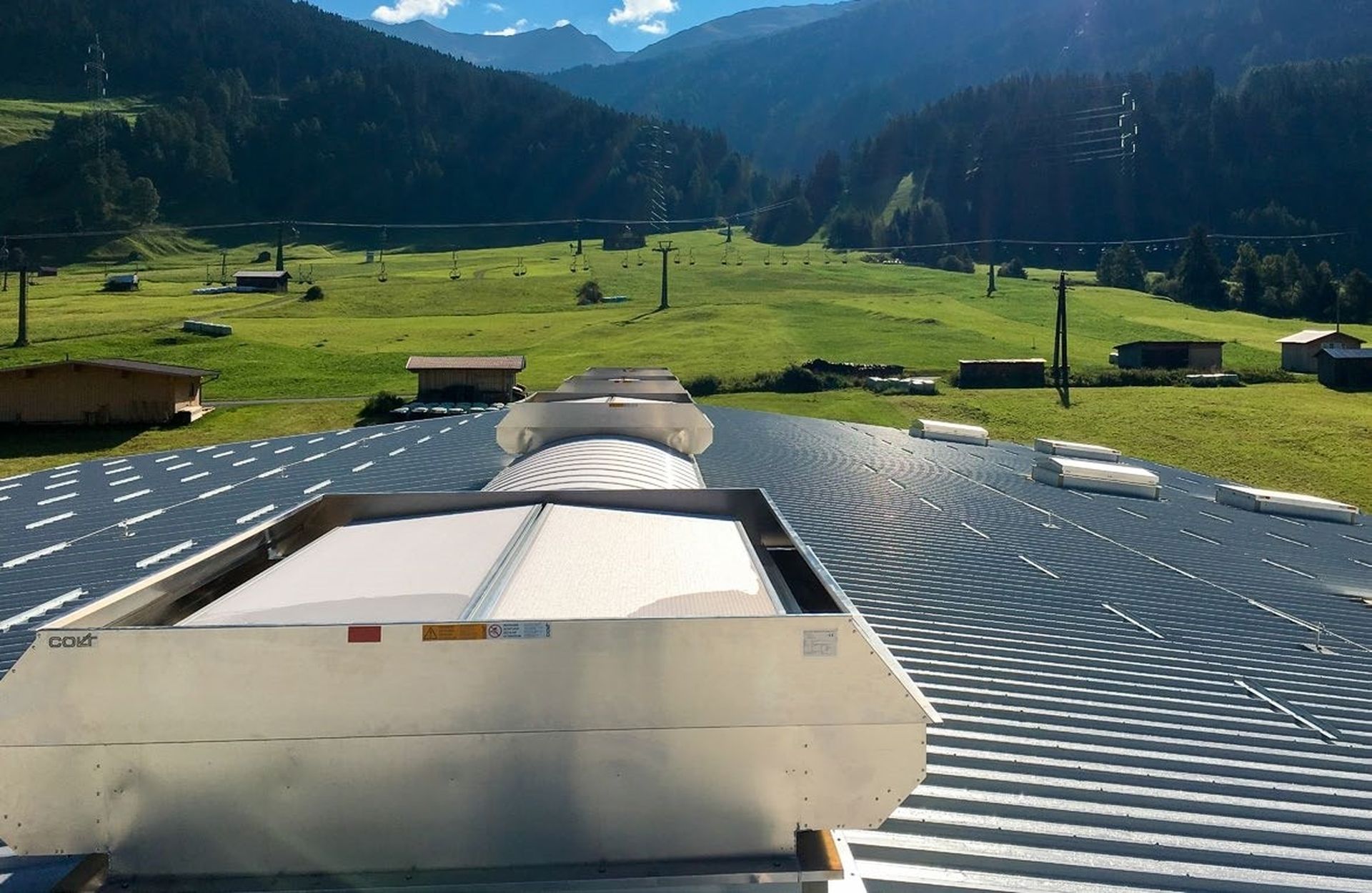 Rooftop vent on a metal roof overlooking a scenic landscape with green fields, scattered buildings, and mountains in the background.
