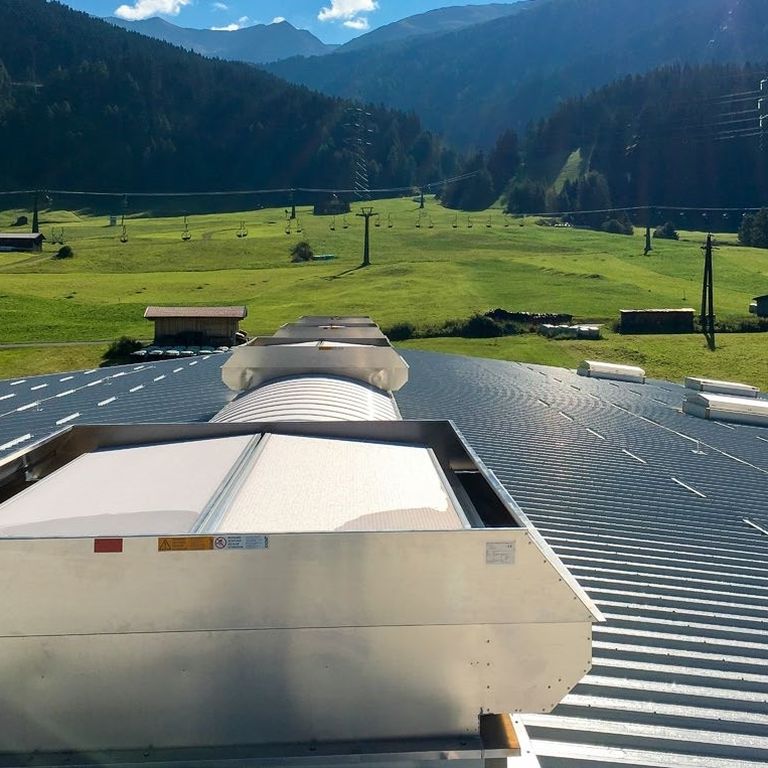 Rooftop vent on a metal roof overlooking a scenic landscape with green fields, scattered buildings, and mountains in the background.