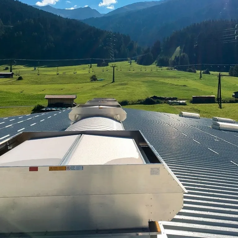 Rooftop vent on a metal roof overlooking a scenic landscape with green fields, scattered buildings, and mountains in the background.