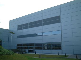 A modern, gray metal-clad building with large windows, set against a cloudy sky, surrounded by a manicured lawn and small shrubs.