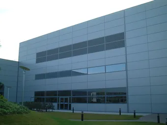 A modern, gray metal-clad building with large windows, set against a cloudy sky, surrounded by a manicured lawn and small shrubs.