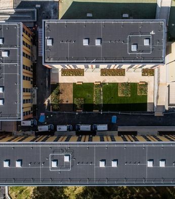 Aerial view of three rectangular buildings with flat roofs, grass courtyards, and parked vehicles along the perimeter in an urban setting.