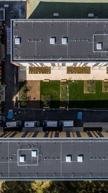 Aerial view of three rectangular buildings with flat roofs, grass courtyards, and parked vehicles along the perimeter in an urban setting.