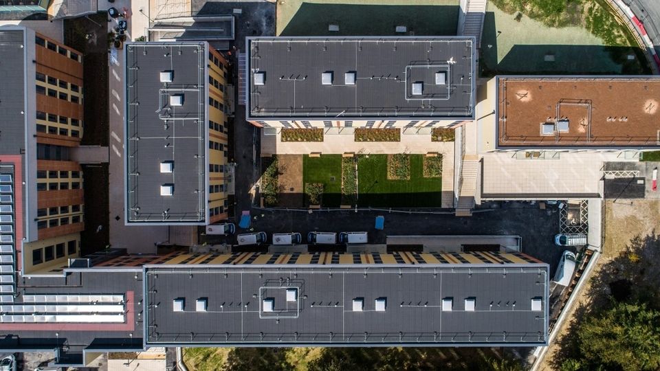 Aerial view of three rectangular buildings with flat roofs, grass courtyards, and parked vehicles along the perimeter in an urban setting.