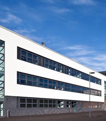 A modern three-story building with large windows and a brick and white facade under a clear blue sky. People are visible inside.