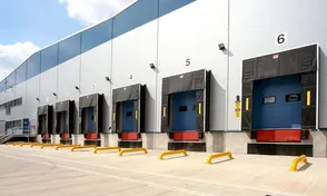 Loading docks with numbered bays lined up on a warehouse exterior, featuring yellow safety barriers and blue doors.