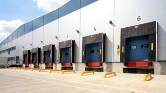 Loading docks with numbered bays lined up on a warehouse exterior, featuring yellow safety barriers and blue doors.