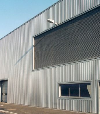 Exterior of a large industrial warehouse with corrugated metal walls, large shutters, few small windows, and an overcast sky.
