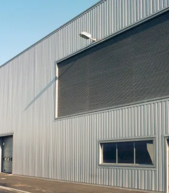 Exterior of a large industrial warehouse with corrugated metal walls, large shutters, few small windows, and an overcast sky.