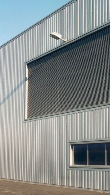 Exterior of a large industrial warehouse with corrugated metal walls, large shutters, few small windows, and an overcast sky.