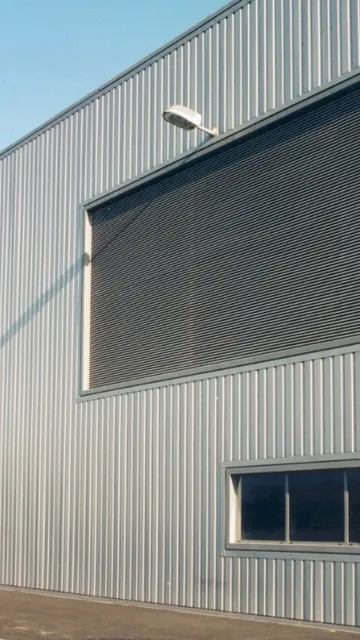 Exterior of a large industrial warehouse with corrugated metal walls, large shutters, few small windows, and an overcast sky.