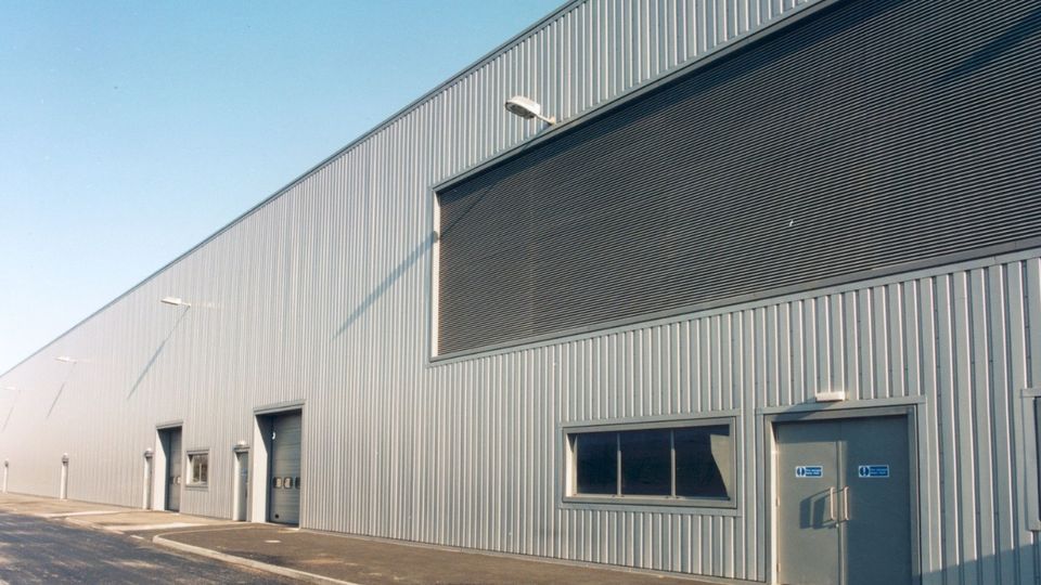 Exterior of a large industrial warehouse with corrugated metal walls, large shutters, few small windows, and an overcast sky.