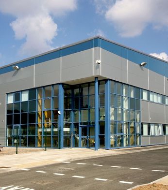 Modern industrial warehouse with large blue-framed windows and a parking area in front, set under a partly cloudy sky.
