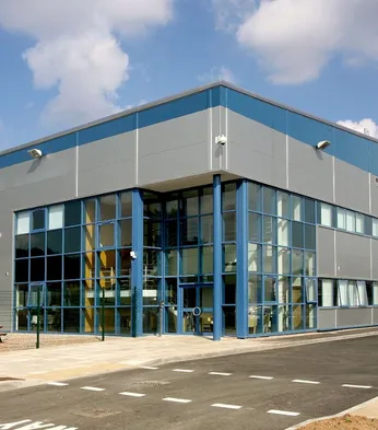Modern industrial warehouse with large blue-framed windows and a parking area in front, set under a partly cloudy sky.