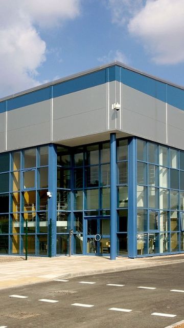 Modern industrial warehouse with large blue-framed windows and a parking area in front, set under a partly cloudy sky.