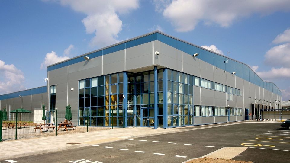 Modern industrial warehouse with large blue-framed windows and a parking area in front, set under a partly cloudy sky.