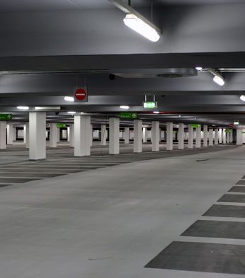 Empty underground carpark with pillars, overhead lights, and marked parking spaces. Signs indicate exit directions.