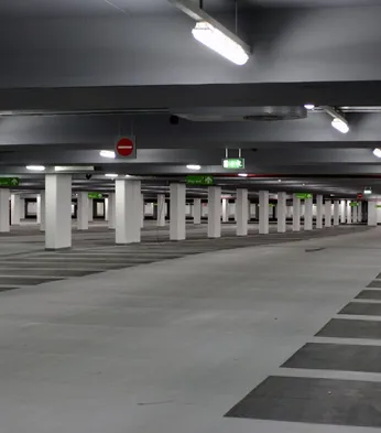 Empty underground carpark with pillars, overhead lights, and marked parking spaces. Signs indicate exit directions.