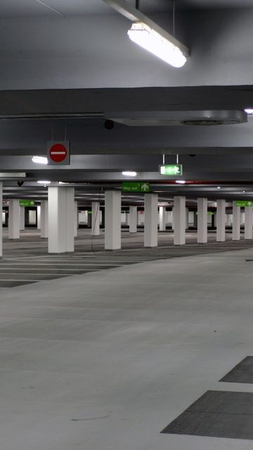Empty underground carpark with pillars, overhead lights, and marked parking spaces. Signs indicate exit directions.