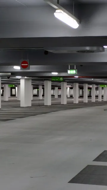 Empty underground carpark with pillars, overhead lights, and marked parking spaces. Signs indicate exit directions.