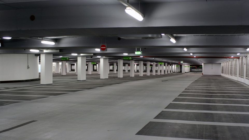Empty underground carpark with pillars, overhead lights, and marked parking spaces. Signs indicate exit directions.