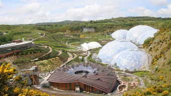 Aerial view of the Eden Project, a botanical garden with large biome structures, surrounded by greenery and winding paths under a partly cloudy sky.
