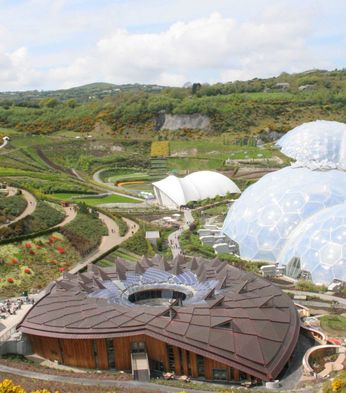 Aerial view of the Eden Project, a botanical garden with large biome structures, surrounded by greenery and winding paths under a partly cloudy sky.