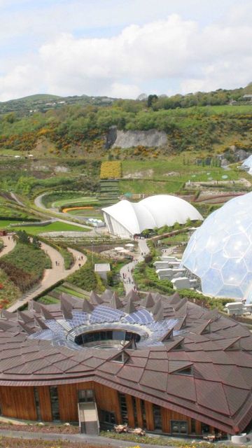 Aerial view of the Eden Project, a botanical garden with large biome structures, surrounded by greenery and winding paths under a partly cloudy sky.