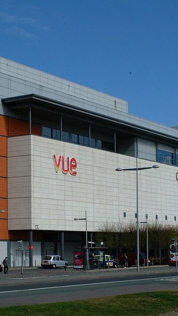 Modern shopping center with a cinema, featuring a mix of white and orange panels, large glass windows, and a few vehicles parked outside.