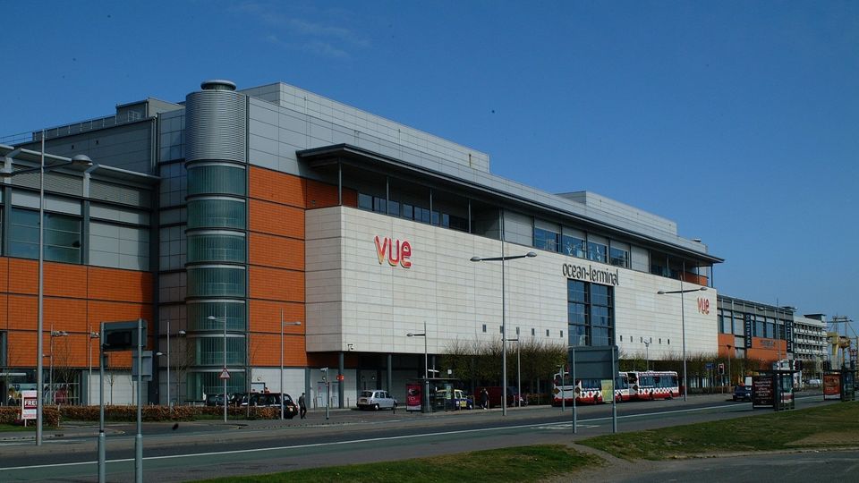 Modern shopping center with a cinema, featuring a mix of white and orange panels, large glass windows, and a few vehicles parked outside.