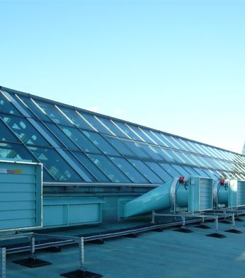 Rooftop view showcasing a slanted glass facade and a large dome skylight with visible smoke ventilator systems under a clear blue sky.