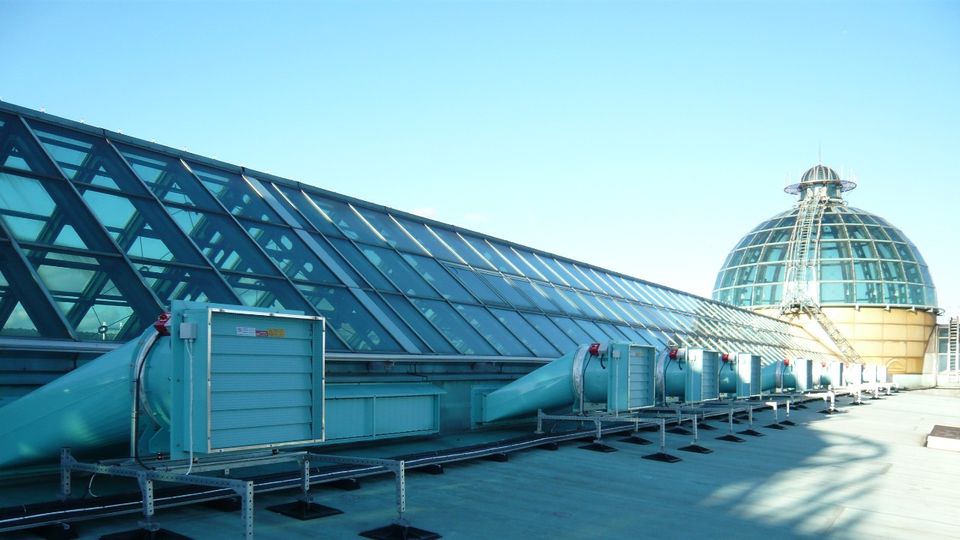 Rooftop view showcasing a slanted glass facade and a large dome skylight with visible smoke ventilator systems under a clear blue sky.