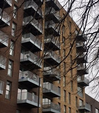 A modern apartment building with multiple balconies, built from brown and tan bricks, partially obscured by bare tree branches.