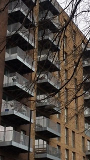 A modern apartment building with multiple balconies, built from brown and tan bricks, partially obscured by bare tree branches.