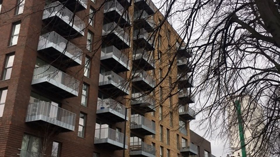 A modern apartment building with multiple balconies, built from brown and tan bricks, partially obscured by bare tree branches.