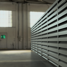 Interior of an industrial warehouse with large metal louvered panels on the right and bright windows in the background.